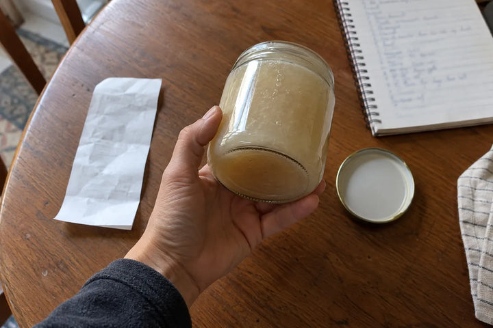 A hand inspecting a plain sea moss gel jar beside its lid and a shopping notebook